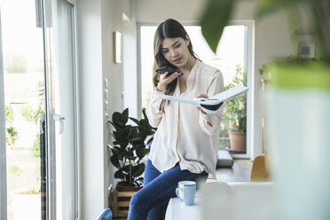 Young woman sitting at table at home holding smartphone and plane model - UUF16955