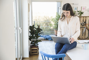 Junge Frau sitzt zu Hause am Tisch und benutzt einen Laptop - UUF16956