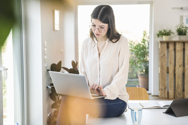 Junge Frau sitzt zu Hause am Tisch und benutzt einen Laptop - UUF16958