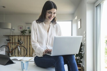 Smiling young woman sitting on table at home using laptop - UUF16960