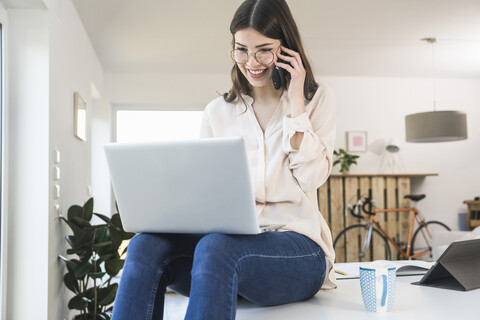 Smiling young woman sitting on table at home using laptop and cell phone stock photo