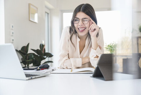 Porträt einer glücklichen jungen Frau, die zu Hause am Tisch sitzt und blinzelt, lizenzfreies Stockfoto