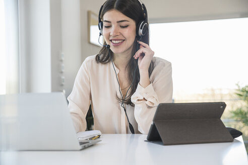 Junge Frau sitzt zu Hause am Tisch und trägt ein Headset - UUF16972