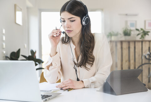 Young woman sitting at table at home wearing a headset - UUF16974