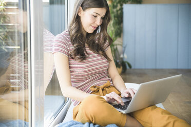 Young woman sitting at the window at home using laptop - UUF16976