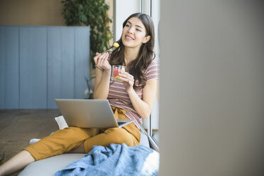 Junge Frau sitzt zu Hause am Fenster mit Laptop und isst eine Kleinigkeit - UUF16978