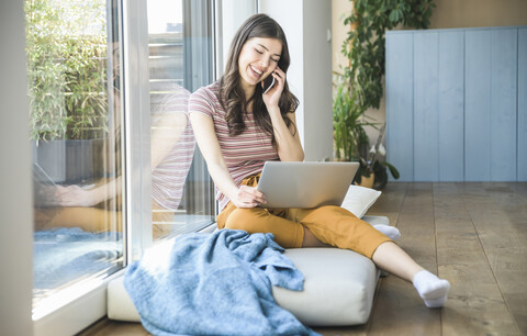 Glückliche junge Frau sitzt zu Hause am Fenster und benutzt Handy und Laptop, lizenzfreies Stockfoto