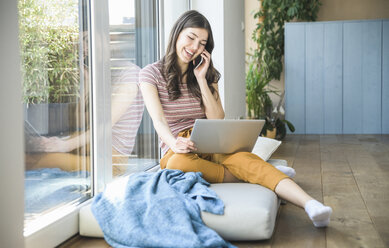 Happy young woman sitting at the window at home using cell phone and laptop - UUF16979