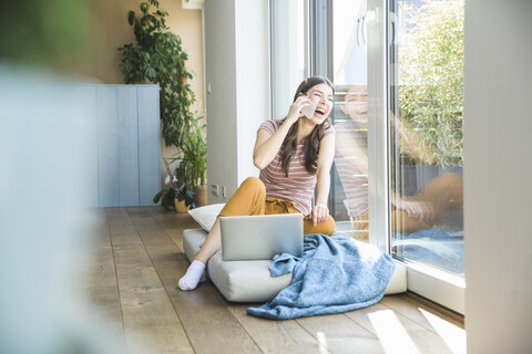 Lachende junge Frau, die zu Hause am Fenster sitzt und Handy und Laptop benutzt, lizenzfreies Stockfoto