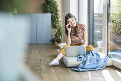 Lächelnde junge Frau sitzt zu Hause am Fenster und benutzt Handy und Laptop - UUF16981