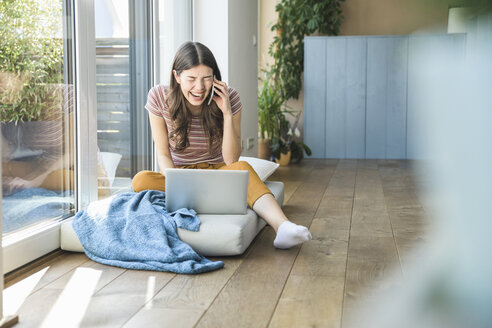 Lachende junge Frau, die zu Hause am Fenster sitzt und Handy und Laptop benutzt - UUF16982