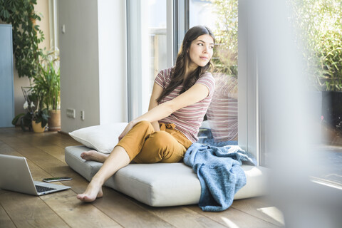 Junge Frau sitzt am Fenster zu Hause mit Laptop, lizenzfreies Stockfoto