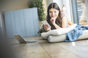 Porträt einer lächelnden jungen Frau mit Mobiltelefon, die zu Hause am Fenster liegt - UUF16988