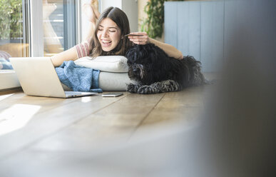 Happy young woman with dog lying at the window at home shopping online - UUF16992