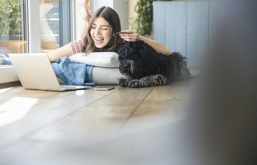 Glückliche junge Frau mit Hund am Fenster liegend beim Online-Shopping zu Hause - UUF16992