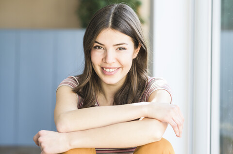 Porträt einer lächelnden jungen Frau, die zu Hause am Fenster sitzt, lizenzfreies Stockfoto