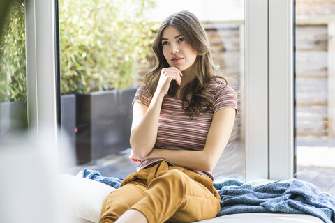 Pensive young woman sitting at the window at home stock photo