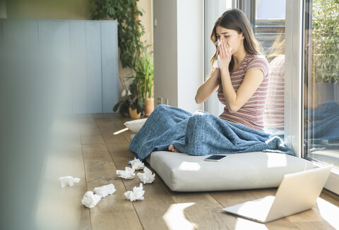 Junge Frau, die zu Hause am Fenster sitzt und sich mit ihrem Laptop die Nase putzt, lizenzfreies Stockfoto