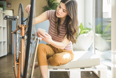 Junge Frau mit Mobiltelefon repariert Fahrrad zu Hause, lizenzfreies Stockfoto