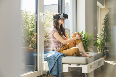 Junge Frau sitzt am Fenster zu Hause und trägt eine VR-Brille, lizenzfreies Stockfoto