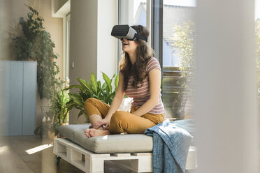 Happy young woman sitting at the window at home wearing VR glasses - UUF17020