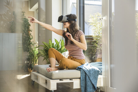 Junge Frau sitzt am Fenster zu Hause und trägt eine VR-Brille, lizenzfreies Stockfoto