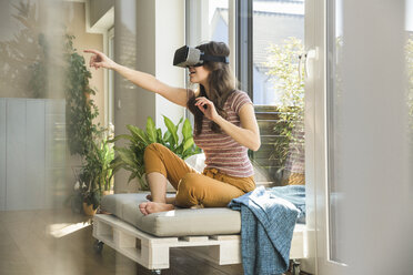 Junge Frau sitzt am Fenster zu Hause und trägt eine VR-Brille - UUF17023