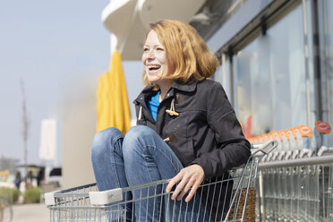 Portrait of laughing woman sitting in shopping cart in front of supermarket - SGF02351