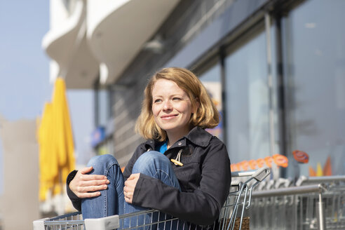 Porträt einer lächelnden Frau, die in einem Einkaufswagen vor einem Supermarkt sitzt - SGF02352