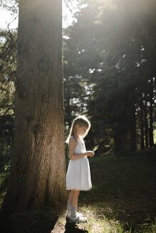 Kleines Mädchen in weißem Kleid steht an einem Baum - EYAF00100