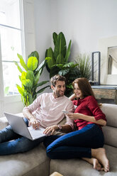 Mann und Frau sitzen auf der Couch, arbeiten am Laptop und benutzen ein Smartphone - GIOF06117