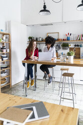 Two women working together in kitchen, using laptop, discussing documents - GIOF06127