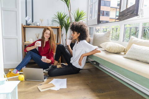 Lässige Geschäftsfrauen sitzen auf dem Boden, trinken Kaffee und arbeiten am Laptop, lizenzfreies Stockfoto