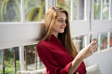 Beautiful woman sitting on bench at the window, using smartphone - GIOF06149