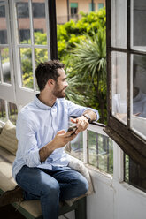 Junger Mann sitzt auf einer Bank am Fenster und benutzt sein Smartphone - GIOF06156