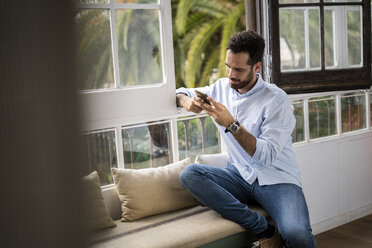 Junger Mann sitzt auf einer Bank am Fenster und benutzt sein Smartphone - GIOF06157
