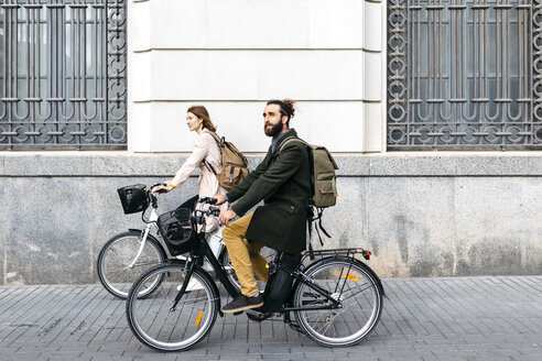Ein Paar fährt mit dem E-Bike in der Stadt an einem Gebäude vorbei - JRFF02901