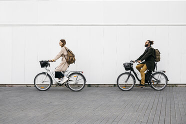 Couple riding e-bikes along a white wall in the city - JRFF02908