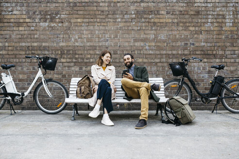 Glückliches Paar, das auf einer Bank neben E-Bikes sitzt und sein Mobiltelefon teilt - JRFF02926