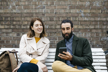 Couple sitting on a bench at a brick wall sharing cell phone - JRFF02927