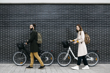 Couple pushing e-bikes along a brick wall - JRFF02952