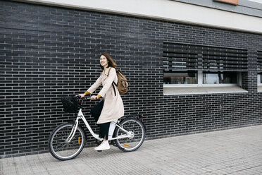 Woman riding e-bike along a brick wall in the city - JRFF02955