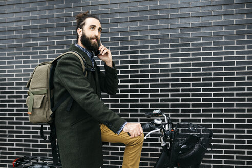 Man with e-bike talking on cell phone at a brick wall - JRFF02956