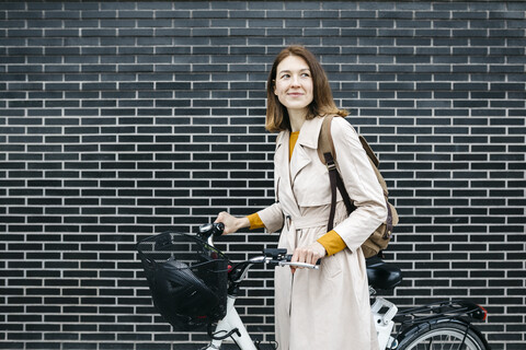 Lächelnde Frau mit E-Bike an einer Backsteinmauer, lizenzfreies Stockfoto