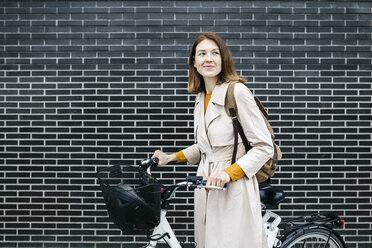 Smiling woman with e-bike at a brick wall - JRFF02963