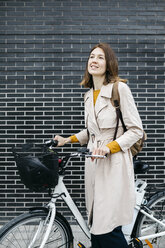 Smiling woman with e-bike at a brick wall - JRFF02964