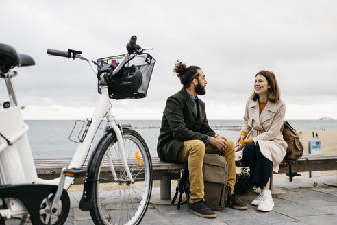 Paar sitzt auf einer Bank an der Strandpromenade neben einem E-Bike und unterhält sich - JRFF02966