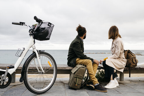 Paar sitzt auf einer Bank an der Strandpromenade neben einem E-Bike und schaut auf das Meer - JRFF02968