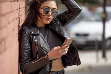 Porträt einer jungen Frau mit Sonnenbrille und schwarzer Lederjacke, die auf ihr Smartphone schaut - OCMF00377