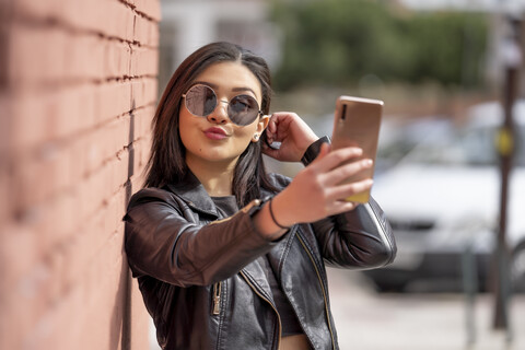 Porträt einer jungen Frau mit Sonnenbrille und schwarzer Lederjacke, die ein Selfie mit ihrem Smartphone macht, lizenzfreies Stockfoto